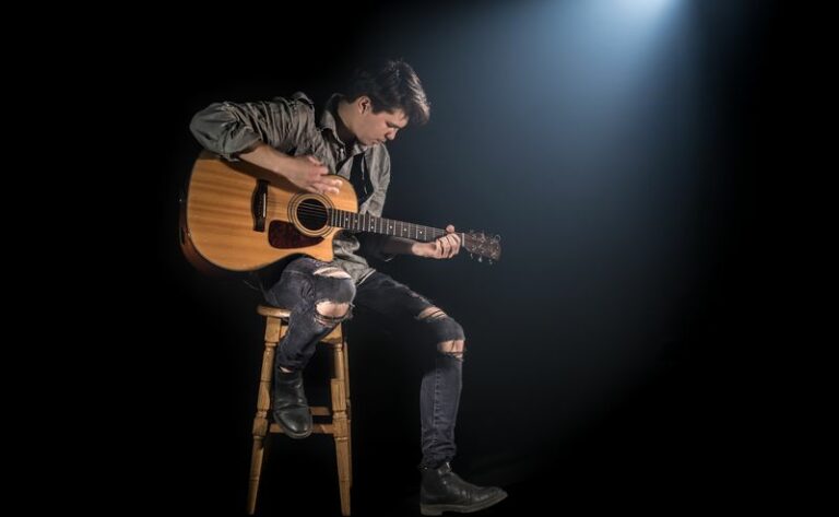 musician playing acoustic guitar, sitting on high chair, black background with beautiful soft light